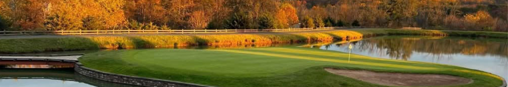 Scenic golf course with a green, water features, and autumn foliage in the background.