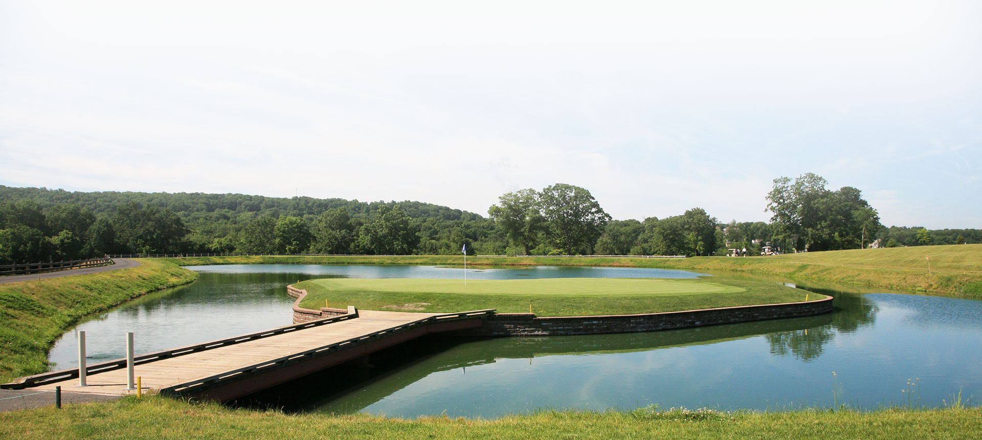 A scenic view of a golf course with a pond, wooden bridge, and lush greenery under a clear sky.