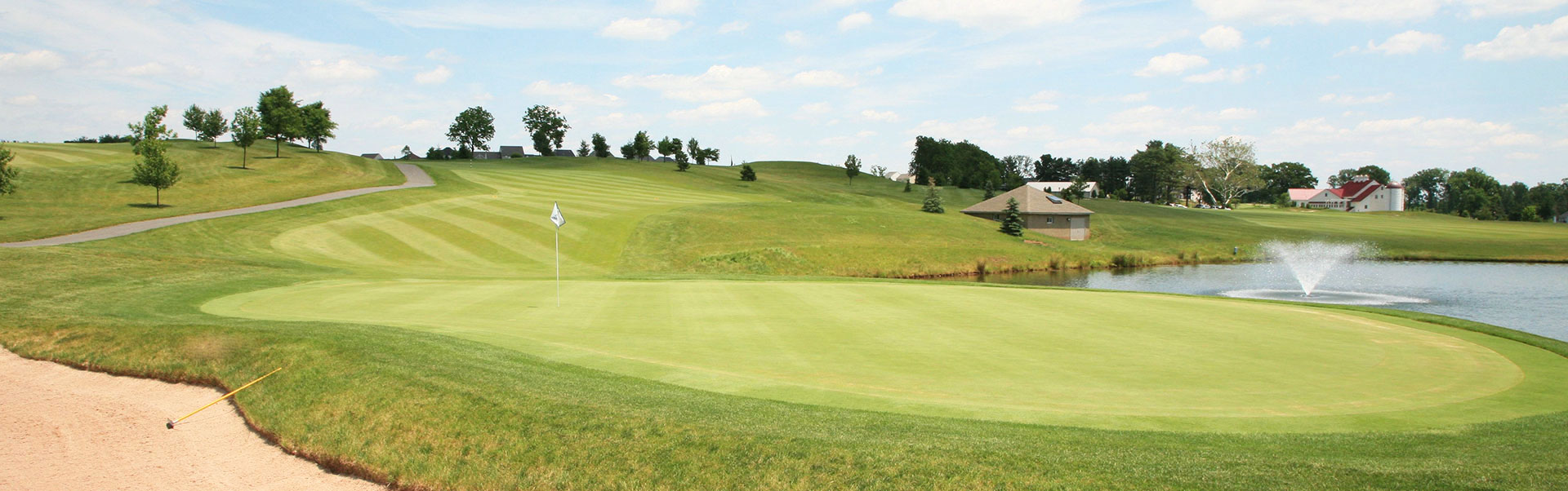 A scenic golf course with a green, sand trap, and a pond featuring a fountain under a blue sky.