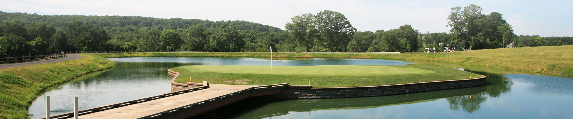 A scenic view of a golf course with a pond, green grass, and trees in the background under a clear sky.